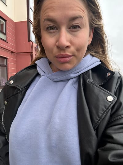A medium shot photo, taken from a slightly low angle, shows a fair-skinned woman with light brown hair and green eyes, looking directly at the camera. She is wearing a light blue hooded sweatshirt and a black leather jacket. Her mouth is slightly open. Behind her, a building with pink and cream-colored walls and dark windows is visible. The sky is overcast. The image appears to have been taken outdoors. The location is identified as Veliky Novgorod, Russia, though no specific landmarks are identifiable to confirm this.