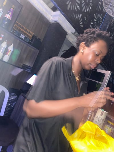 A woman is standing behind a counter in a dimly lit establishment, possibly a bar or club, in Howayi, Nigeria. She is wearing a black collared shirt and has dark, short, curly hair. She is leaning over the counter, looking down at something, and her hands are blurred as she appears to be in motion. On the counter in front of her, there are various items including a bottle of Teeling Whiskey, a shaker, and a bright yellow plastic bag. Behind her, shelves are visible containing bottles and other objects. The environment appears to be a commercial setting with a patterned light fixture on the ceiling and a fan overhead.