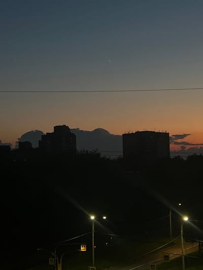 This image captures a twilight scene in Shushary, Russia. Silhouettes of apartment buildings rise against a dusky sky, with clouds tinged by the setting sun. Below, streetlights illuminate a roadway, casting a warm glow. Pedestrian crossing signs are visible, indicating urban infrastructure. The overall atmosphere is one of quiet transition from day to night.