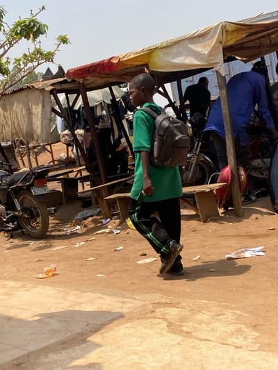 The image portrays an outdoor street or market scene in Katcha, Nigeria, under clear daylight. A young male, appearing to be a boy, walks from left to right across the mid-foreground. He wears a green short-sleeved shirt, black athletic pants with two green stripes along the outer seams, and dark shoes, carrying a dark grey backpack over both shoulders. He holds a small white object in his right hand and looks slightly towards his right.

The ground surface is predominantly reddish-brown earth, with scattered debris including plastic and paper. A lighter-colored, possibly concrete or tiled, surface occupies the immediate foreground. In the background, multiple makeshift market stalls are visible. One prominent stall features a wooden frame supporting a yellow and orange tarp roof, with white fabric sections, from which clothes appear to be hanging. To the left, a black motorcycle with a Nigerian license plate "KEE-133NG" is parked. Further right and deeper in the scene, blurred figures are visible, including one person in a blue garment leaning over another motorcycle, and another individual in black. Overhead, a clear, light blue sky is visible, with sparse tree branches and green leaves extending from the upper left. Shadows are cast on the ground, consistent with direct sunlight.