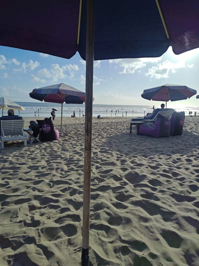 The image depicts a beach scene in Kuta, Indonesia, on a sunny day. The foreground is dominated by textured sand, with shadows cast by undulating dunes. In the mid-ground, beach umbrellas provide shade. On the left, a person sits in a white lounge chair facing the ocean, with a black beanbag chair beside them. Further back, another umbrella shades a low table and two purple beanbag chairs, occupied by a person in a lounge chair. Several other beach umbrellas and beanbag chairs are visible throughout the scene. In the background, the ocean waves gently roll onto the shore, and numerous people can be seen walking, swimming, or relaxing. The sky is a clear blue with scattered white clouds, and the sun is shining brightly, casting a warm glow over the entire scene. The overall impression is one of a vibrant and relaxed beach atmosphere.