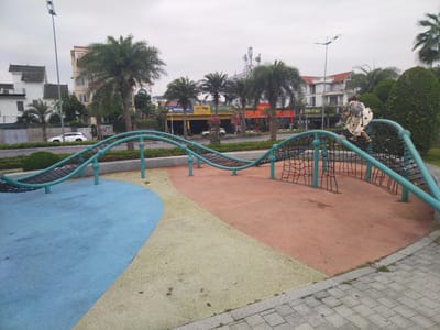 The image depicts a playground in the city of Ha Long Ward, Vietnam. A young girl in a light-colored, polka-dotted dress and sandals is climbing on a play structure featuring teal-colored support poles and a black net bridge. The playground surface is divided into sections of blue, yellow, and reddish-brown material.
In the background, palm trees line a street with various businesses on the opposite side, including an establishment with "MICHE" visible on a yellow sign, and others with red signs bearing Vietnamese script. Buildings of various architectural styles, some with red-tiled roofs, can also be seen. The sky is overcast and gray. A white car is parked in front of the street. Paved areas are visible, bordering the playground.