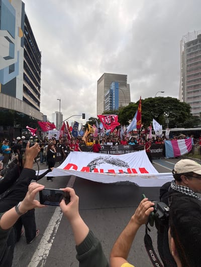 The image captures a protest or demonstration taking place on a city street. A large group of people are marching, holding flags and a large banner. The banner has text and a graphic on it. People are taking photos and videos of the protest. The setting appears to be an urban environment, with tall buildings on either side of the street. The location is São Paulo, Brazil. The sky is overcast.