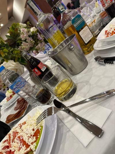 The image depicts a table setting, likely in Elhovo, Bulgaria, as indicated by the Cyrillic writing on a water bottle reading "Орна Баня," a brand of bottled water. The table is covered with a white tablecloth and cluttered with a variety of items. There are multiple bottles of alcohol, including a bottle of Bushmills 10-year-old Irish whiskey. There is also a bottle of Coke and a bottle of water. Some of the bottles are lying down and some are standing up. There is an ice bucket, likely for keeping drinks cool. Cutlery, including a fork and a spoon, is placed on a napkin. Plates containing food, one with salad topped with grated cheese and another with sliced meat, are also visible. A corkscrew is also present. A floral arrangement with white and pink flowers adds a decorative touch to the table. The scene suggests a festive occasion with food and drinks.