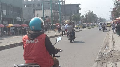 A motorcyclist in a blue helmet and red vest with "TVHQL" on the back rides on a busy street. Other motorcycles and vehicles share the road. Pedestrians walk on the right side. Shops and buildings line the left side of the street, some shaded by red umbrellas. The weather appears sunny.