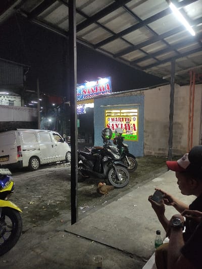 This nighttime image captures a street-side scene at Warteg Sanjaya in Bogor, Indonesia. A large blue and white "SANJAYA" sign illuminates the background, situated above a light-blue building with a prominent yellow "WARTEG SANJAYA" sign on its facade. The immediate foreground is covered by a corrugated metal roof supported by black metal pillars, lit by overhead fluorescent tubes.
To the right, a person wearing a red and white baseball cap and a dark shirt is seated, holding a smartphone horizontally with both hands, focused on its screen. A watch is visible on their left wrist. A second individual's arm is partially visible to the left of the first person. On the concrete ground near the seated figures, a clear plastic cup and a green plastic bottle are visible, along with scattered cigarette butts.
In the middle ground, two black motor scooters (one identifiable as a Honda Beat) are parked, facing left, each with a helmet hanging from its handlebars. A brown chicken is on the concrete ground near these parked vehicles. To the left, a white panel van is parked facing right. The front wheel and partial yellow and blue fender of another motorbike are visible in the extreme left foreground. The concrete ground appears worn, with visible dirt and some moist patches. A darker, possibly industrial building is present in the distant left background.