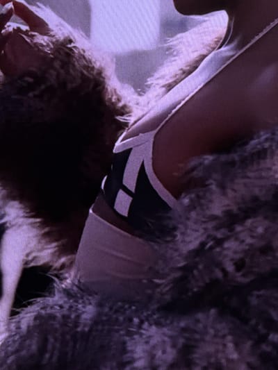 The image displays a close-up view of a person's shoulder and upper torso, suggesting an indoor setting. The person is wearing a light-colored garment with a dark, geometric design on the strap. A furry, textured material, possibly a scarf or boa, is draped around the shoulder, obscuring parts of the background. The lighting appears to be soft, casting a purple hue over the scene. There is no identifiable information to pinpoint a specific location within Dubai, United Arab Emirates, or any indication of specific actions or interactions.