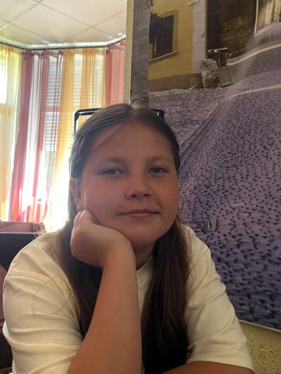 A young female subject, appearing to be in her pre-teen years, is depicted indoors in a medium close-up, located in Novopokrovskaya, Russia. She has long, straight, dark brown hair and wears a light cream or off-white short-sleeved t-shirt. Dark-framed eyeglasses are perched on top of her head. She faces the camera directly with a neutral expression, resting her chin on her right hand, which is supported by an unseen surface.
The background is visually divided. To the subject's left, a window is covered by multi-colored sheer curtains in shades of yellow, orange, and reddish-pink, allowing diffused natural light to enter. A white grid-patterned acoustic tile ceiling is visible in the upper left. To the subject's right, a wall is adorned with a large mural or tapestry depicting an antiquated street scene. Features of the mural include a yellow building with a dark, recessed window and a textured, cobblestone-like ground rendered in shades of grey and purple. The scene suggests a casual indoor setting.