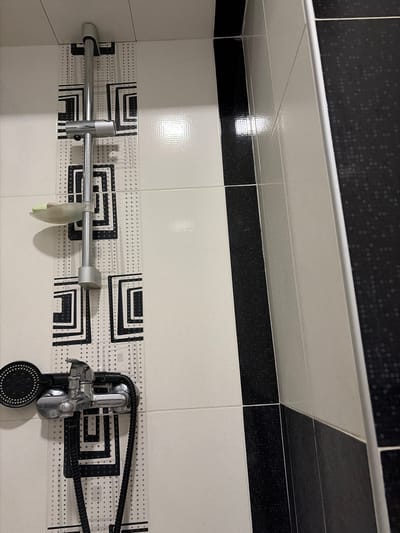 The image shows a close-up of a shower fixture and tiled wall in a bathroom. The shower fixture is made of chrome and includes a faucet, showerhead, and a hose. Above the faucet, there is a soap dish. The wall is tiled with a black and white geometric pattern. The lighting suggests an indoor environment, likely a bathroom. There are no people present in the image. The specific location in Sliven, Bulgaria, is not discernible from the visual information provided.