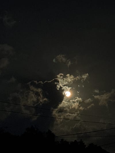 The image depicts a nighttime scene in Tinaquillo, Venezuela. The moon is visible through scattered clouds, with a bright glow emanating from its position in the sky. Three electrical power lines diagonally cut through the sky. The dark silhouettes of trees appear on the right-hand side of the image. At the left, a very dim light indicates the presence of another object.