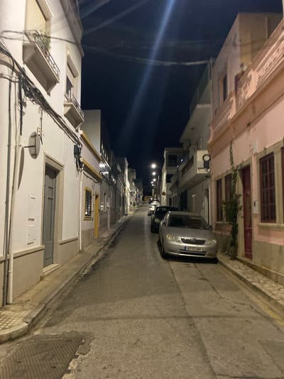 The image shows a narrow street in Olhão, Portugal, at night. The street is lined with buildings on both sides, some of which are lit. There are several cars parked on the street, and a street light is visible in the distance. The overall atmosphere is quiet and peaceful.
