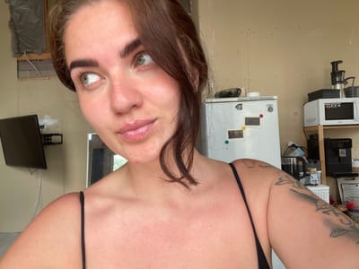A young woman with brown hair and green eyes is posing for a selfie in what appears to be a construction or unfinished room. She is wearing a black spaghetti strap top. The background includes a white refrigerator with magnets on it, a microwave on a shelf, a coffee machine, a black computer monitor mounted on the wall, and some construction materials like insulation and drywall. The room has unpainted walls and exposed framing in some areas. The woman is looking off to the side with a slight smile.