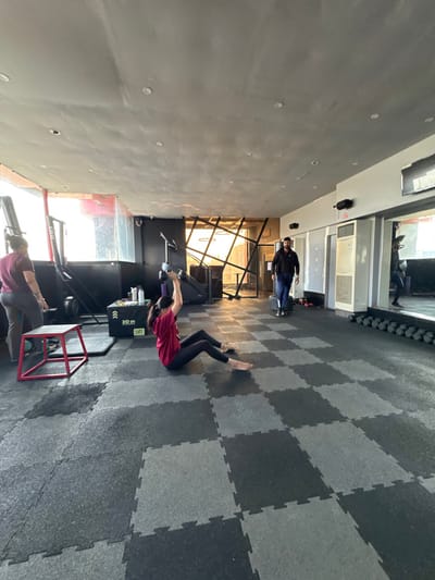 A gym features checkered floors and light gray walls with recessed lighting. A woman in a maroon shirt and black leggings performs sit-ups near a black box with yellow lettering. Nearby, another person in gray pants and a maroon top is engaged in a different exercise using gym equipment. A third individual, dressed in dark clothing, stands in the background near windows. The gym's interior design includes exposed support structures in the far wall.