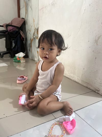 A young child sits on a tiled floor, playing with toys. The child is wearing a white tank top and patterned shorts. In front of the child are two pink toy houses, a bracelet made of colorful beads, and a pink shoe. A box of tissues and a stroller are visible in the background. The room appears to be in a home located in Kecamatan Pati, Indonesia. The walls are a neutral color with some discoloration and scuff marks.