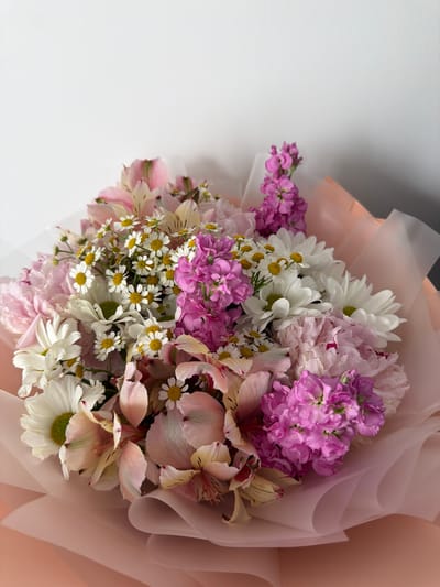 A close-up, eye-level studio shot features a bouquet of various flowers, predominantly in shades of pink, white, and pale yellow, with accents of vibrant purple. The flowers are arranged in a rounded, dense fashion and are partially wrapped in sheer pink paper. The background is a plain, light gray wall, with a shadow visible on the right side. The lighting is bright and even. There are no people or animals present. The bouquet is the sole focus of the image. The context of Ust'-Dzhigutinka, Russia, is not visually inferable from the provided image.