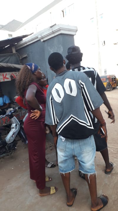 The image shows three people standing outside in Nkpor, Nigeria. Two men are standing close together; the man closest to the camera is wearing a gray jersey with the number 00 on the back, denim shorts, and black sandals. The second man is wearing a black shirt and blue denim shorts. A woman is standing close to the two men, wearing a maroon dress. The ground is concrete. A tricycle can be seen in the background.