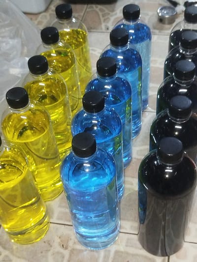 In Si Sunthon, Thailand, a collection of clear plastic bottles filled with colorful liquids sits on a tiled surface. The bottles are capped with black lids. There are three distinct liquid colors: yellow, blue, and a dark, almost black, liquid. The yellow bottles are arranged in a diagonal line from the bottom left towards the center right of the image. The blue bottles are positioned to the right of the yellow ones, also in a diagonal arrangement. The dark liquid is in bottles located on the far right of the image. The lighting suggests an indoor setting, possibly a storage area or a shelf, with reflections visible on the curved surfaces of the bottles. The background is blurred but appears to show more shelving or containers, and a white plastic bag is partially visible in the upper left corner.