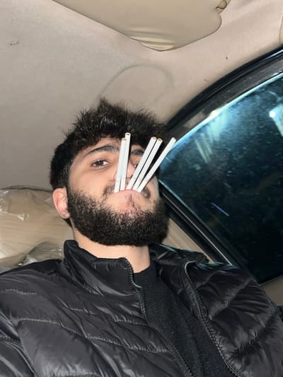 A person with dark hair and a beard is lying down, facing the camera, inside a car. They are wearing a black puffer jacket. Four cigarettes are held in their mouth, extending outwards like whiskers. The car's window and part of the interior are visible in the background. The image is oriented on its side.