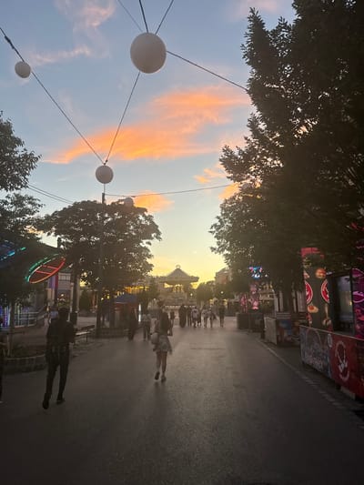 The image depicts an evening scene in the Prater amusement park in Vienna, Austria. The sky displays vibrant sunset colors with orange and pink clouds against a light blue backdrop. Several white, spherical lanterns are suspended by wires, casting a soft glow. The foreground shows an asphalt path lined with trees and various attractions. On the left, there are structures with colorful lights, including a circular neon display. Further down the path, people are walking and socializing. In the distance, a carousel is visible, illuminated and operational. On the right side of the path, there are large advertising banners for food stalls and attractions, such as "FOOD TRACK" and "MAGIC TEN." The overall atmosphere suggests a lively and festive environment in the amusement park as evening approaches.