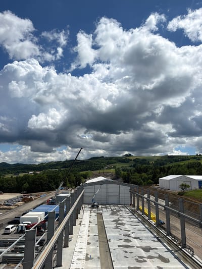 This is a high-angle, eye-level outdoor photograph of a construction site in Stróża, Poland, under a dramatic sky filled with dark, heavy clouds interspersed with patches of blue. The foreground shows the skeletal structure of a building under construction, with large grey steel beams forming vertical supports and horizontal crossbeams. The ground in this area is a concrete slab with numerous water puddles, indicating recent rain.
To the left of the foreground, a construction area is visible with stacks of materials, vehicles including cars, vans, a truck, and a small excavator. Further back, a large white tent-like structure is visible, and beyond that, a crane with its boom extended upwards. In the midground, rolling green hills covered with trees form the landscape, with a few scattered buildings. To the right, another white tent-like structure is situated on a slight incline. The overall scene depicts an active construction project in a rural or semi-rural setting with challenging weather conditions.