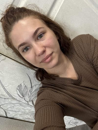 A young woman with fair skin, light-brown hair, and green eyes is pictured in a close-up, tilted headshot. She is wearing a brown ribbed long-sleeve shirt. The background consists of a white surface with a patterned design, possibly a floral or textured wallpaper, and a white door in the distance. There is no indication of the location being Nizhnyaya Gar', Russia. The photo does not provide forensic details or information about actions or interactions.