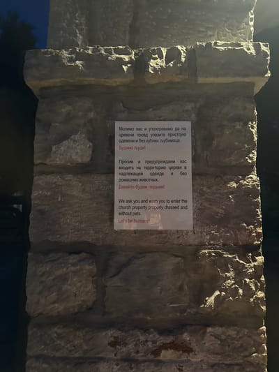 The image shows a close-up view of a stone pillar at night, illuminated by a light source from the left. A white sign is affixed to the pillar, featuring text in both Cyrillic and Latin alphabets. The Cyrillic text reads: "Молимо вас и упозоравамо да на црквени посед улазите пристојно одевени и без кућних љубимаца. Будимо људи! Просим и предупреждаем вас входить на территорию церкви в надлежащей одежде и без домашних животных. Давайте будем людьми!" The Latin text reads: "We ask you and warn you to enter the church properly dressed and without pets. Let's be humans!" The sign appears to be a set of rules for entering a church property, requesting visitors to be appropriately dressed and not bring pets. The stone pillar is made of rough-hewn blocks, with some evidence of weathering. The background is dark, suggesting it is nighttime, with a hint of foliage on the left side. The location is identified as Boreti, Montenegro.