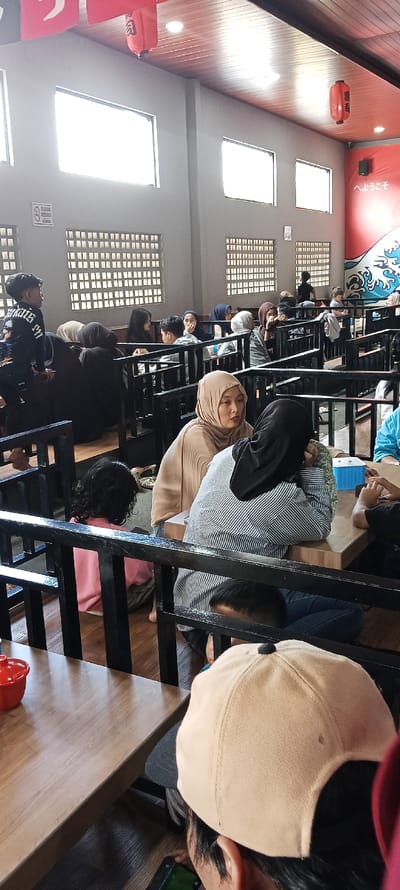 The image depicts a restaurant interior in Katapang, Indonesia, located at coordinates -7.026 latitude, 107.549 longitude. The scene is a typical restaurant setting. Several people are seated at tables, engaging in conversation and consuming food. There is a person seated at a table with a black cap and a red shirt. Another person, wearing a tan hijab, is seated at the same table. A third person is seated across from them, wearing a blue shirt and a black cap. The restaurant is adorned with wooden panels and white walls. Black metal railings separate the tables.