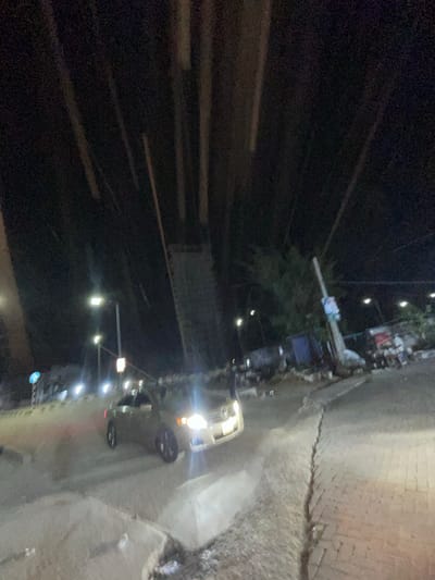 At night in Ilado, Nigeria, a street scene unfolds. The road is wet, reflecting the glare of vehicle headlights. A vehicle with illuminated headlights is parked near the roadside. Streetlights cast a dim glow on the surroundings. One or more pedestrians appear to be in close proximity to the parked vehicle. A sidewalk runs along the edge of the road, lined with buildings. Overhead power lines are visible.
