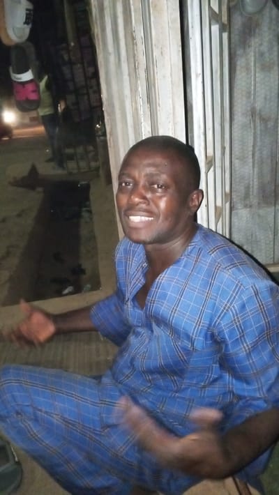 A male individual is depicted in Sabon Tasha, Nigeria, in a low-light outdoor setting. The subject, an adult male with a shaved head and dark complexion, is seated or squatting low to the ground, smiling directly towards the camera. He wears a blue and white plaid two-piece garment, consisting of a long-sleeved tunic with an open collar and matching pants. His hands are slightly blurred, suggesting motion, with his right hand open and raised. To the subject's right, a light-colored, vertically oriented metallic structure, possibly a partially open security gate or metal bars, is visible. To his left, several pairs of sandals, including one white and one black/pink, hang vertically as if on display. The background to the left reveals a dimly lit area with blurred figures of other individuals and what appears to be merchandise on shelves, indicating a storefront or market area. Distant artificial light sources, possibly from streetlights or vehicles, are present further in the background. The ground immediately surrounding the subject is dark and undefined.