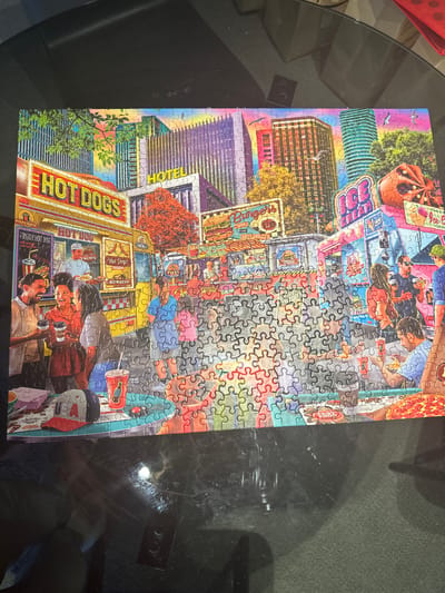 The image depicts a partially completed jigsaw puzzle featuring an urban scene, possibly inspired by a generic cityscape. The puzzle includes a variety of elements: tall buildings with rainbow colored glass facades, hot dog stands with lit signage, and people eating at outdoor tables. Signage includes words such as "HOT DOGS", "TACOS", "BURRITO" and "HOTEL" printed on the jigsaw. The center of the puzzle is incomplete, filled with unassembled pieces that are mainly gray and red. The puzzle sits on a dark, reflective surface. The actual location of the scene is not clearly identifiable, and Clifton, United States of America could be a reference point for the style and character represented in the image, but cannot be forensicly identified based on the jigsaw puzzle on the image.
