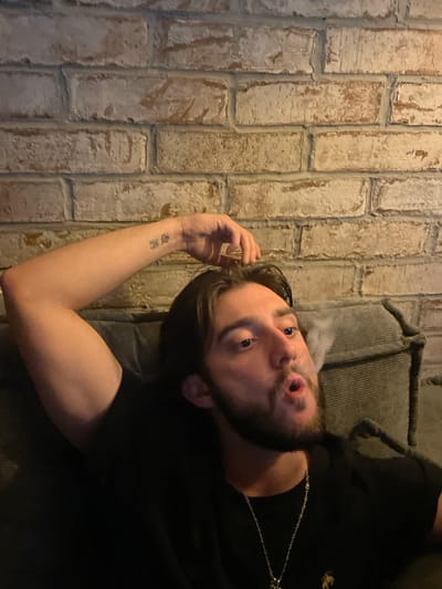 The image shows a man with a beard and dark hair exhaling smoke, possibly from a cigarette or vape, in front of a brick wall. He is wearing a black t-shirt and a necklace. The man is leaning back on what appears to be a couch or armchair. The background is a textured brick wall. There is a tattoo on his left forearm. The lighting is dim, suggesting an indoor setting.