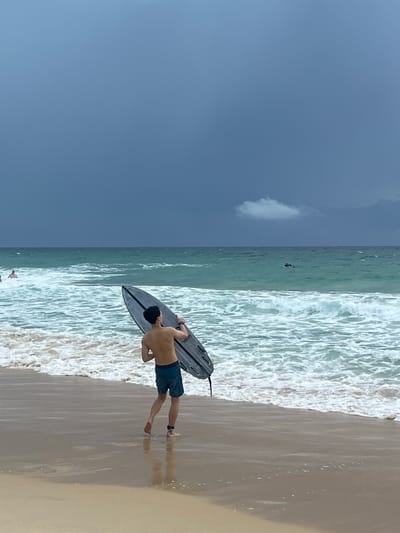 A man, viewed from behind, stands on a sandy beach holding a black bodyboard. He is wearing blue shorts. The man is facing the ocean, with waves crashing onto the shore. The ocean water is a greenish-blue color, and the sky is a deep, overcast blue with a single, small white cloud visible. The overall atmosphere appears to be cloudy, suggesting it might be an overcast day, possibly late afternoon or early morning. The scene is set on a beach, with the wet sand reflecting the sky and the man. In the distance, a few other people are visible in the water, appearing as small figures. The lighting is subdued, consistent with cloudy weather. There is no visible text in the image. The setting is likely Choeng Thale, Thailand, as indicated by the context provided.