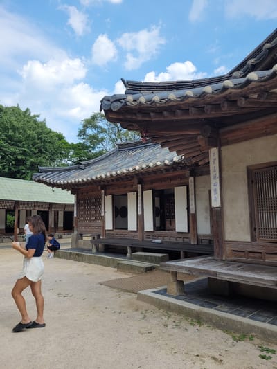 The image depicts a scene at Gyeongbokgung Palace in Seoul, South Korea, under a bright, cloudy sky. In the foreground, a young woman with long, brown hair is standing and looking at her phone. She is wearing a dark blue cropped t-shirt and white shorts, with black slip-on sandals. Behind her, a person wearing a blue jacket and dark pants is crouching on a stone ledge. To the left, another person in a white t-shirt and dark pants stands near a building with a green tiled roof.
The main focus of the image is a traditional Korean building with a dark tiled roof and wooden architecture. The building features intricate wooden latticework on its windows and doors. Several signs with Korean calligraphy are affixed to the building's pillars. A raised wooden platform runs along the front of the building, supported by stone pillars. The ground is covered in light-colored gravel with scattered patches of green vegetation. The background shows lush green trees and the blue sky with scattered white clouds.