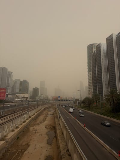 The image depicts an urban landscape under a severe sandstorm or dust storm, casting an ochre-brown hue across the sky and significantly reducing visibility. The scene is located along the Ayalon Highway in Tel Aviv-Yafo, Israel, characterized by its multiple lanes, adjacent railway tracks, and surrounding high-rise buildings.
In the foreground, a concrete barrier separates the viewer's perspective from a dry, unpaved channel with muddy patches and some standing water on the left, running parallel to railway tracks. Two sets of railway tracks with overhead catenary systems are visible on a concrete embankment. Further left, a cluster of modern high-rise buildings, including one with a red billboard displaying Hebrew text, are partially obscured by the dust.
To the right, a multi-lane highway, carrying vehicles in both directions, occupies the midground. Several cars and at least one white van are present on the road; traffic appears moderate. Orange traffic cones are visible along the highway's right edge. A landscaped median strip with palm trees and green foliage separates the highway from a line of prominent, modern high-rise buildings on the far right. One of these buildings, a tall, curved structure, clearly displays "ADGAR" near its top. Other adjacent buildings feature distinctive vertical dark and light paneling. The cityscape extends into the distance, with numerous structures blurred and rendered indistinct by the heavy atmospheric dust. No individuals are visible outside of vehicles.