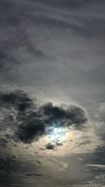 The image is a vertical, low-angle shot of a cloudy sky, captured from Kamensk-Uralsky, Russia. The sky is filled with dense, dark gray clouds, with some lighter, wispy clouds interspersed. The sun is partially obscured by a large, dark cloud mass in the lower center of the frame, creating a bright, hazy glow around its edges. The sun appears as a bright white disc with a halo effect due to the surrounding clouds. There are no visible people, animals, or man-made structures. The scene appears to be a natural outdoor environment, likely during daytime. A very small dark speck, possibly a bird or an insect, is visible in the upper portion of the sky.