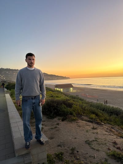 A man stands on a tiled overlook at a beach. He wears jeans, a grey sweater, and dark sneakers. He is holding something in his hand. Behind him, a coastal scene unfolds: a beach with a lifeguard station, ocean waves, and a horizon with a gradient sunset. Vegetation grows on a sandy hillside adjacent to the overlook. A distant cityscape is visible along the coastline.