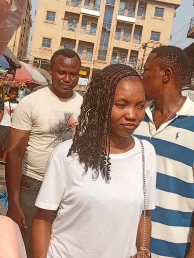 The image displays an outdoor street or market scene in Fegge, Nigeria, under bright daylight. A young adult female, with black braided and curly hair, occupies the foreground, wearing a plain white short-sleeved t-shirt and a silver-chained bag strap over her right shoulder. She is positioned slightly angled to the right, looking in that direction with a neutral expression. Behind her, to the left, stands an adult male with short dark hair, wearing a light-colored t-shirt featuring a dark graphic design; he gazes downwards. To the right and slightly behind the foreground female, an adult male with short dark hair and a blue and white horizontally striped polo shirt (with a visible Ralph Lauren logo) has his head partially obscured by the foreground female, looking towards the left. A thin gold-colored chain is visible around his neck. In the mid-ground to the far left, a child or young adolescent female in a white t-shirt with a graphic stands near market stalls, identifiable by red and white striped umbrellas and hanging produce, possibly dried onions or garlic. The background features a prominent multi-story building, light tan in color, equipped with multiple windows and balconies featuring metallic railings. Overhead utility wires and poles are visible between the subjects and the background building.