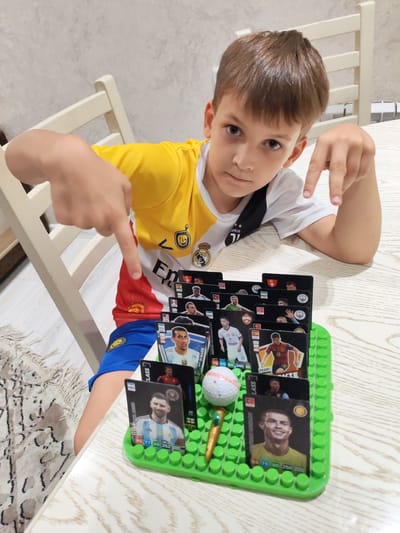 A young boy with brown hair, wearing a yellow and white shirt with football club logos, is sitting at a wooden table and pointing at a collection of football player trading cards displayed on a green plastic holder. The cards feature images of various footballers, including Lionel Messi and Cristiano Ronaldo. A golf ball and a pen-like object are also on the holder. The boy is looking at the camera with a serious expression. The background shows a white chair and a light-colored wall, with a rug on the floor. The scene appears to be indoors, in a domestic setting. There is no visible indication that this scene is located in Tashkent, Uzbekistan.