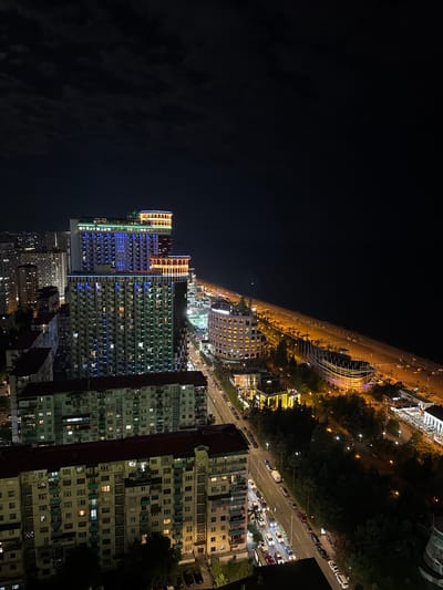 The image is an aerial night view of a cityscape, likely Batumi, Georgia, showcasing a blend of modern and older architecture. A multi-lane road, illuminated by streetlights, runs through the scene, with rows of cars parked along its sides and some driving. Trees line the road, creating a green contrast against the urban environment.

Dominating the foreground are several tall buildings. Some are brightly lit with colorful LED lights, featuring orange and blue hues, giving them a vibrant and modern appearance. These buildings have numerous windows, many of which are illuminated, suggesting residential or office spaces. Adjacent to these are older, more densely built apartment blocks, characterized by their repetitive window patterns and weathered facades. One building in the foreground is partially obscured by a darker structure.

In the background, a long elevated structure, possibly a highway or promenade, stretches across the horizon, its supports and pathway clearly visible. Beyond this, the cityscape continues, with more buildings and lights receding into the darkness. The sky above is a deep black, indicating it is nighttime. The lighting from the buildings and streetlights creates a warm, inviting glow across the urban landscape. No people are distinctly visible, suggesting the activity captured is the general urban life at night, with the focus being on the illuminated architecture and infrastructure.