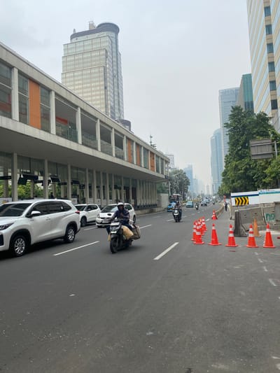 The image depicts a street scene in Central Jakarta, Indonesia, on an overcast day. A multi-lane road is visible, with white cars and a scooter in motion. On the left side of the road, there is a modern building with a covered walkway and orange accents. In the background, tall skyscrapers dominate the skyline, indicative of a city center. The right side of the road shows ongoing construction work, with orange traffic cones and concrete barriers lining a section of the road. A few pedestrians are also present, along with more vehicles further down the road. The overall impression is of a busy urban environment experiencing development.