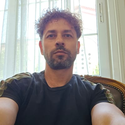 A man with curly brown hair and a beard is pictured from the chest up, seated in a carved wooden chair. He is wearing a black t-shirt with a camouflage pattern on the sleeves. The background consists of a sheer white curtain and a window with vertical blinds, indicating an indoor setting with natural light. There is no specific identifiable landmark of Athens, Greece, in the image. The scene is static and focuses on the portrait of the man.