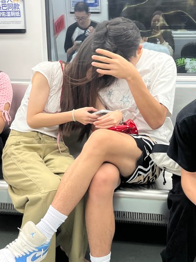 In Shapingba District, China, two individuals are seated side-by-side on a public transportation seat, possibly a subway or train. The person on the left, appearing to be female, wears a white eyelet top and tan cargo pants. She is holding a mobile phone in her hands, with a red polka dot clutch bag resting on her lap. A light green bracelet adorns her left wrist. The person on the right, appearing to be male, is wearing a white t-shirt with star embellishments and black shorts with striped accents. His right arm is raised, covering his face, and his legs are crossed over the other individual's legs. Both are seated on a metallic bench with a ribbed ventilation grate beneath. In the background, reflections in a window show other passengers, including a man in a black and white shirt and glasses. A poster with Chinese text is visible on the left wall.