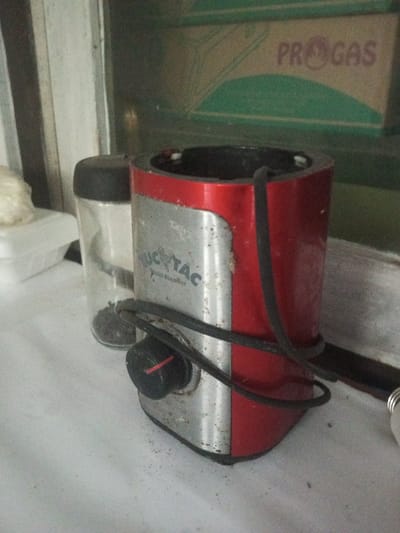 The scene, located in Ciherang, Indonesia, displays a still life arrangement on a white, speckled surface. Dominating the center is a red and silver "TUC TAC Mini Blender" base. The blender features a silver-toned front panel inscribed with "TUC TAC Mini Blender," flanked by vertical red panels. A black control knob with a red indicator mark is affixed to the lower front. A black electrical cord is loosely wrapped around the lower section of the appliance. To the left of the blender, a transparent cylindrical spice grinder with a black cap holds dark, granular material. A portion of a white styrofoam container is partially visible further to the left. The background features a greenish vertical surface, against which a light brown cardboard box displaying the word "PROGAS" and a green semicircular emblem is present. The white surface supporting the objects shows scattered dark particulate matter. No individuals are visible, and no active interactions are depicted.