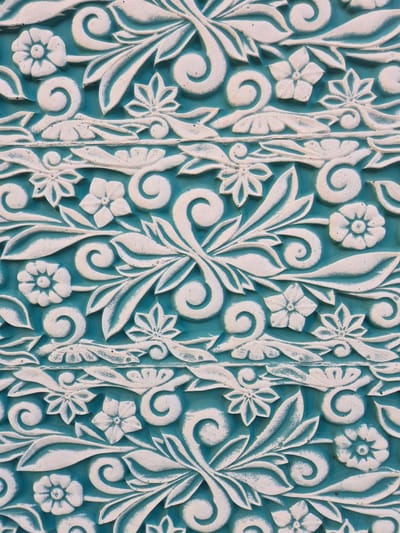 This is a close-up, full-frame shot of a decorative, carved pattern on a surface. The carving is in relief, with a light-colored, possibly white, design set against a teal background. The pattern features stylized floral motifs and swirling, foliate elements. Small, star-shaped flowers with four or five petals are interspersed with larger, elongated leaves and coiled tendrils. The carving has a textured, slightly weathered appearance, with subtle variations in color and shadow highlighting the raised details. Vertical elements resembling reeds or stems run through the pattern, dividing sections of the design. There are no people or animals visible, and the image focuses solely on the intricate details of the decorative carving. The lighting appears even, suggesting it was taken under diffuse conditions, perhaps indoors or on an overcast day. No text is visible in the image. The context provided indicates the image is from Voronezh, Russia, suggesting this carving might be part of architectural ornamentation or a decorative piece within that location.