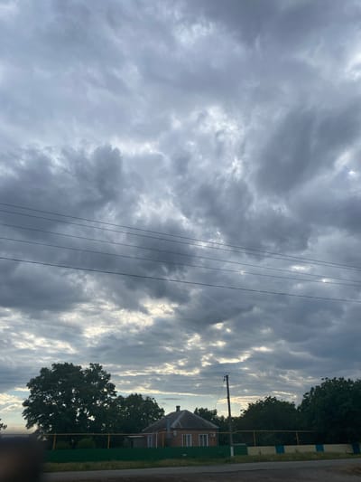 The image captures a landscape scene dominated by a dramatic, cloudy sky. The clouds are a mix of dark, brooding grays and lighter, brighter patches where the sun attempts to break through, creating a dynamic interplay of light and shadow. Several dark power lines stretch diagonally across the frame, cutting through the sky and adding a man-made element to the natural scene.
In the lower right portion of the image, a portion of a building with a tiled roof and brick walls is visible, suggesting a rural or suburban setting. Adjacent to the building are trees with dark green foliage. A fence, partially visible, with yellow and blue markings separates the property from a road. The lighting suggests it is either early morning or late afternoon, given the low angle of the sun implied by the bright patches in the clouds and the overall mood. The weather appears to be overcast, with a potential for rain or recent rain, given the dark clouds. There is no text visible in the image. The scene evokes a sense of quiet, possibly a slightly melancholic or contemplative atmosphere due to the heavy cloud cover.