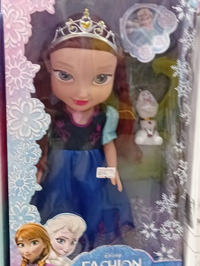 The image shows a doll in a box, likely a toy for sale. The doll is styled after a character from the Disney movie Frozen, with a tiara, blue dress, and braided hair. It is accompanied by a small figure of Olaf, the snowman character from the same movie. The box itself is decorated with snowflake motifs and the Disney logo. A price tag is visible on the doll's dress. The scene appears to be a retail display in a store. The background is blurred, but suggests a commercial environment. The image appears to be taken in Baan Lamai, Thailand, though no specific landmarks are visible.