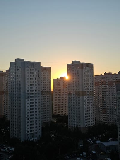 The image shows a view of residential buildings in the city of Odintsovo, Russia, at sunset. The sky is a clear gradient of light blue and yellow, with the sun partially visible between two tall apartment buildings. These buildings are characteristic of Soviet-era architecture, with numerous balconies and windows. In the foreground, the scene transitions to darkness, revealing the tops of trees and a parking lot filled with cars. The lighting suggests that the photograph was taken during the golden hour.