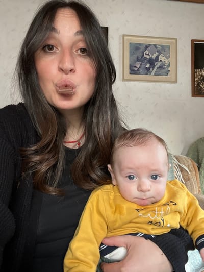 This image depicts an adult female holding an infant in an indoor setting within Wüstenrot, Germany.

The female, positioned centrally and partially cropped at the waist, has long dark hair and wears a black long-sleeved top and a black cardigan. A red beaded necklace is visible around her neck. Her lips are pursed, holding a small, light-brown, rectangular object, possibly a piece of chocolate or candy, and she gazes directly forward.

Cradled in her arms is an infant, appearing to be several months old, with light brown hair and blue eyes. The infant wears a yellow long-sleeved shirt featuring the text "mommy's little boy," partially visible dark-striped pants, and white socks. A distinct wet stain is present on the left side of the infant's yellow shirt. The infant also looks directly forward with a neutral expression.

The background consists of a light-colored wall with a faint, textured pattern. On the wall, to the right of the female's head, hangs a framed aerial photograph depicting residential houses and surrounding terrain, set in a light wooden frame. A second, darker-framed item is partially visible on the far right. Part of an upholstered furniture piece, featuring a woven pattern in brown and green tones, is visible behind the infant on the right side of the frame.