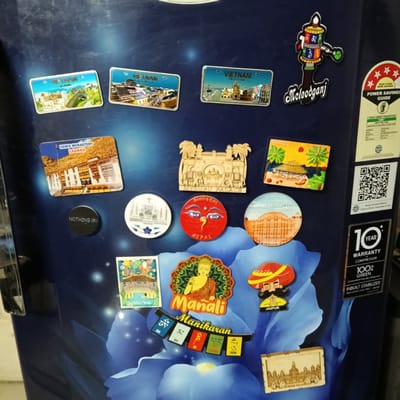 The image shows a refrigerator door covered in various magnets. The magnets depict locations and themes from across Asia, including Vietnam, Ladakh, Nepal, Kerala, and Jaipur. There are also magnets related to Buddhist themes and places like Manali and Manikaran. The refrigerator itself has a blue floral design and includes informational stickers regarding energy efficiency and warranty. The location in Bengaluru, India, is not evident from the image content.