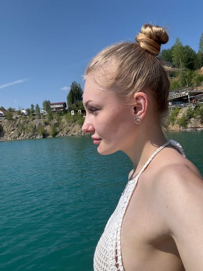 A young Caucasian woman with blonde hair pulled into a bun is shown in profile, facing left. She is wearing a white crocheted halter top and a circular earring in her left ear. She is positioned near a body of turquoise water. In the background, across the water, is a rocky embankment with trees, buildings, and what appears to be a pier with some vehicles. The sky is clear and blue. The scene is likely Karyer Izvestnyak, Russia, known for its former quarry turned into a lake.
